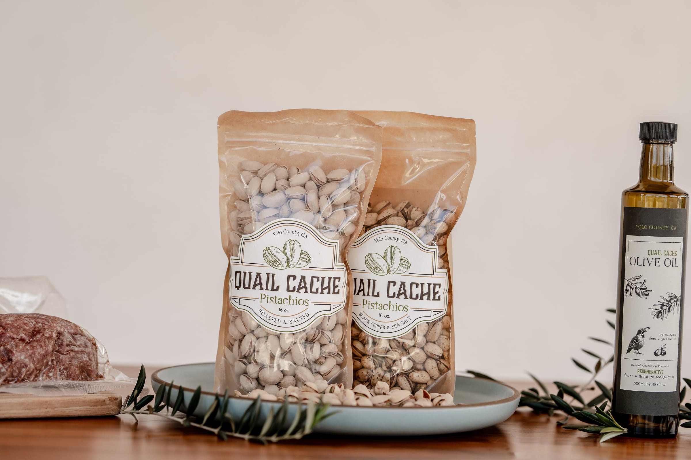 Two bags of Quail Cache pistachios, Roasted & Salted and Black Pepper & Sea Salt, on a plate beside a bottle of the ranch’s olive oil